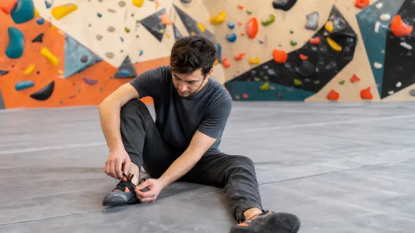 Wspinacz zakłada but wspinaczkowy na materacu przed kolorową ścianą boulderową